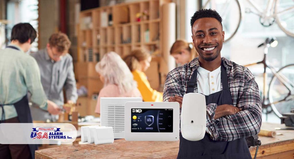 Smiling man in a commercial setting holding a security system device, featuring USA Alarm Systems branding, with coworkers engaged in work in the background, illustrating expert security solutions for businesses in Santa Clarita Valley.
