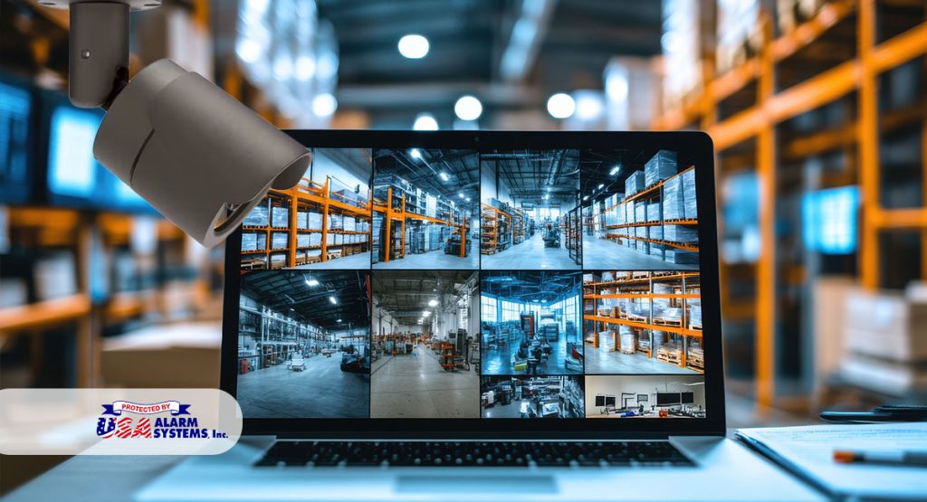 Laptop displaying multiple security camera feeds of a warehouse, with a security camera mounted above, showcasing inventory protection solutions by USA Alarm Systems in a professional setting.